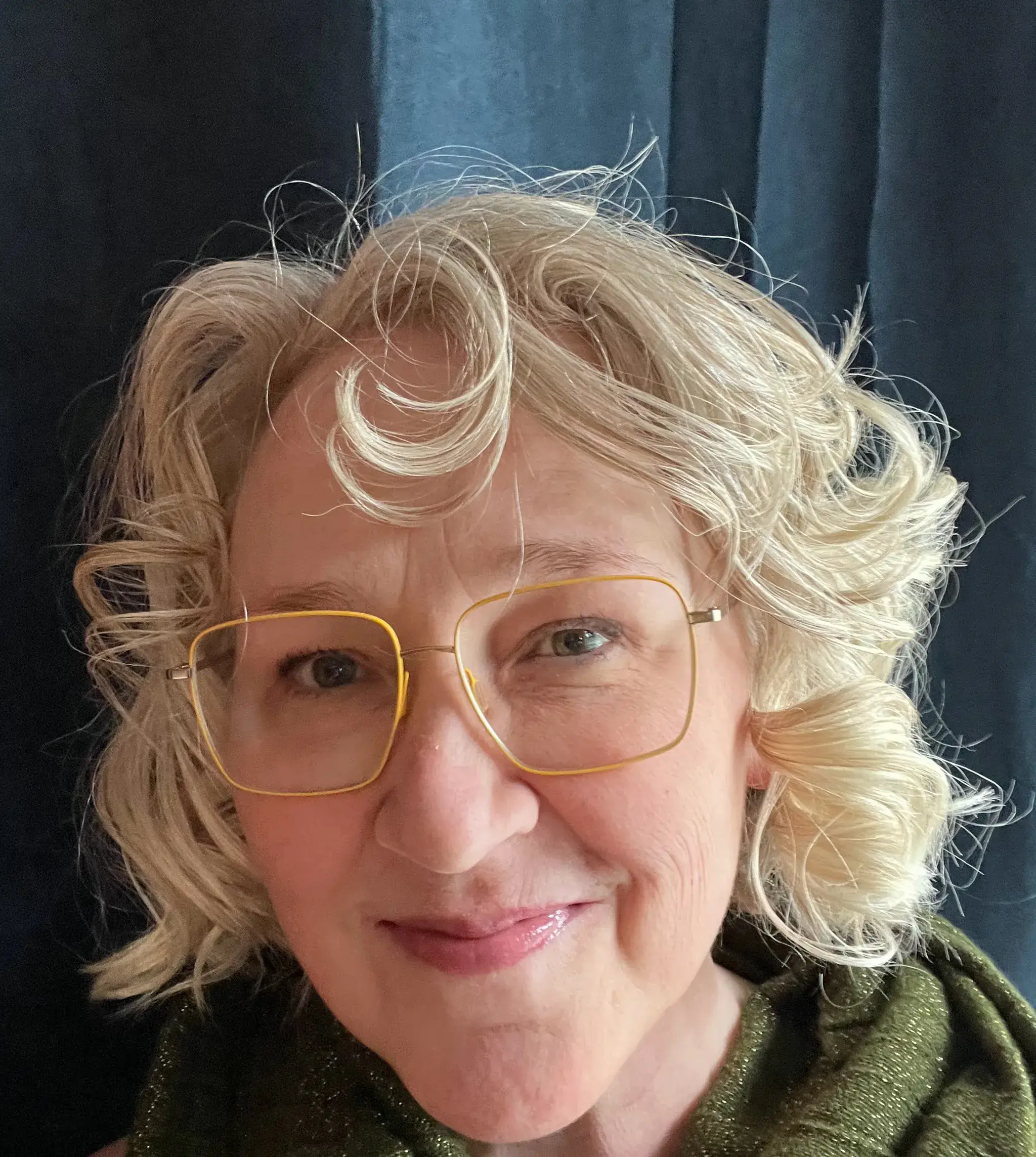 Photo is a headshot of Lisa who appears blonde with curls and is wearing yellow-rimmed glasses. She is in front of a blue background, smiling, and wearing a green scarf.